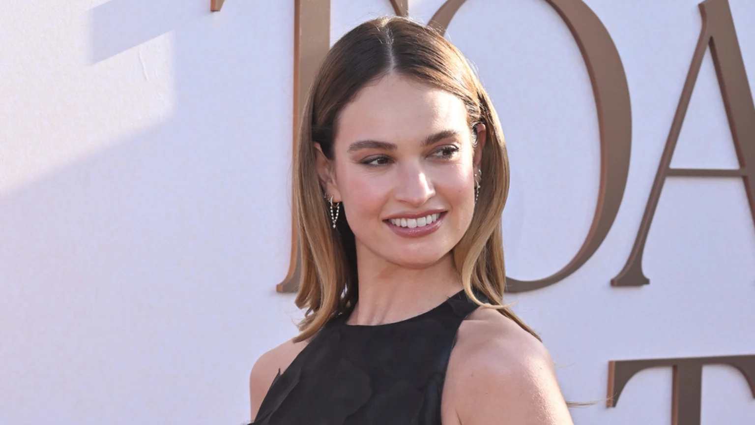 Lily James smiles, looking to the right, with light brown hair, wearing a black top and small dangling earrings against a white wall with large bronze letters in the background.
