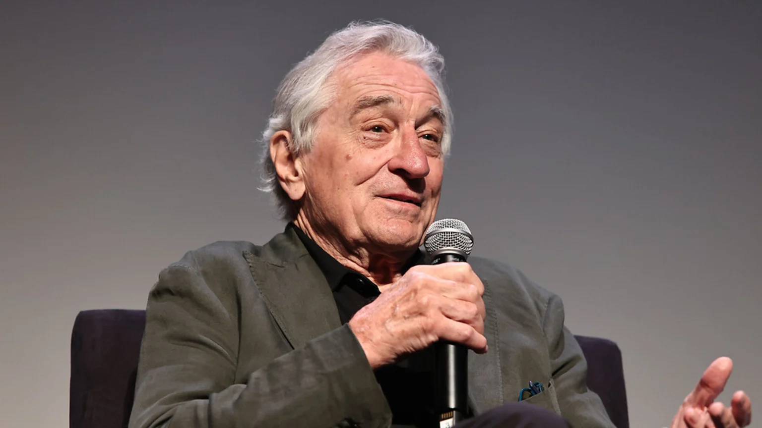 Robert De Niro, an older man with grey hair, sits facing right and holds a microphone to his mouth, wearing a dark jacket and shirt.
