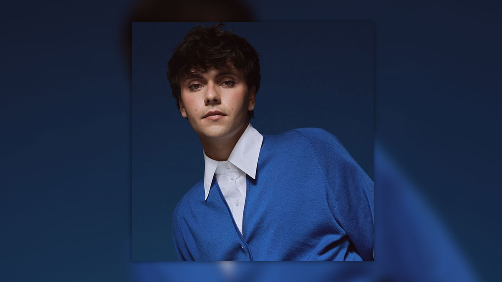 A young person with dark curly hair and subtle eye makeup, wearing a white collared shirt and blue cardigan, looks at the camera against a dark blue background.