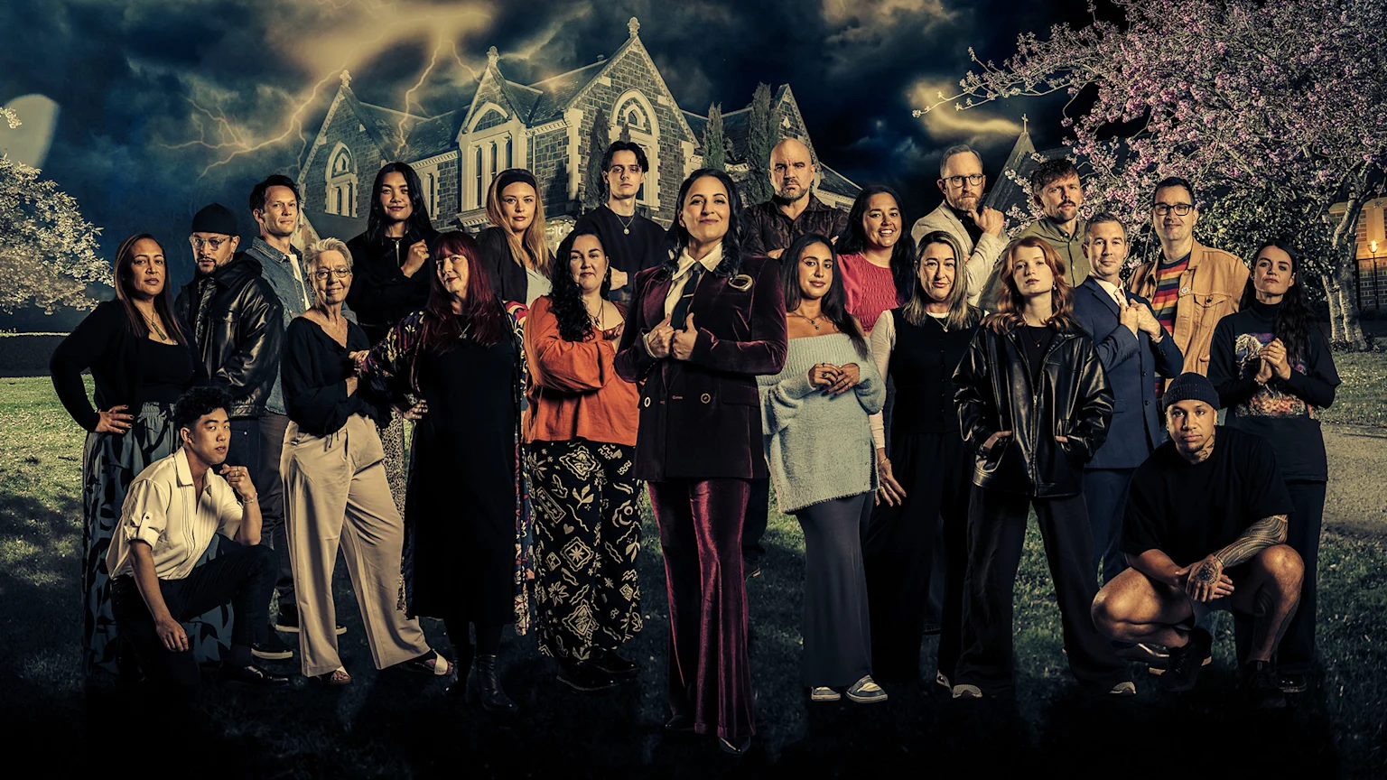 A diverse group of 22 individuals, including the host in a burgundy velvet suit, pose dramatically before a dark mansion under a lightning-filled sky.