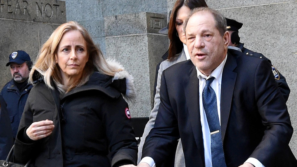 Harvey Weinstein, in a suit and tie, walks with a blonde woman in a black parka, accompanied by other people and a stone building inscribed "FEAR NOT" in the background.