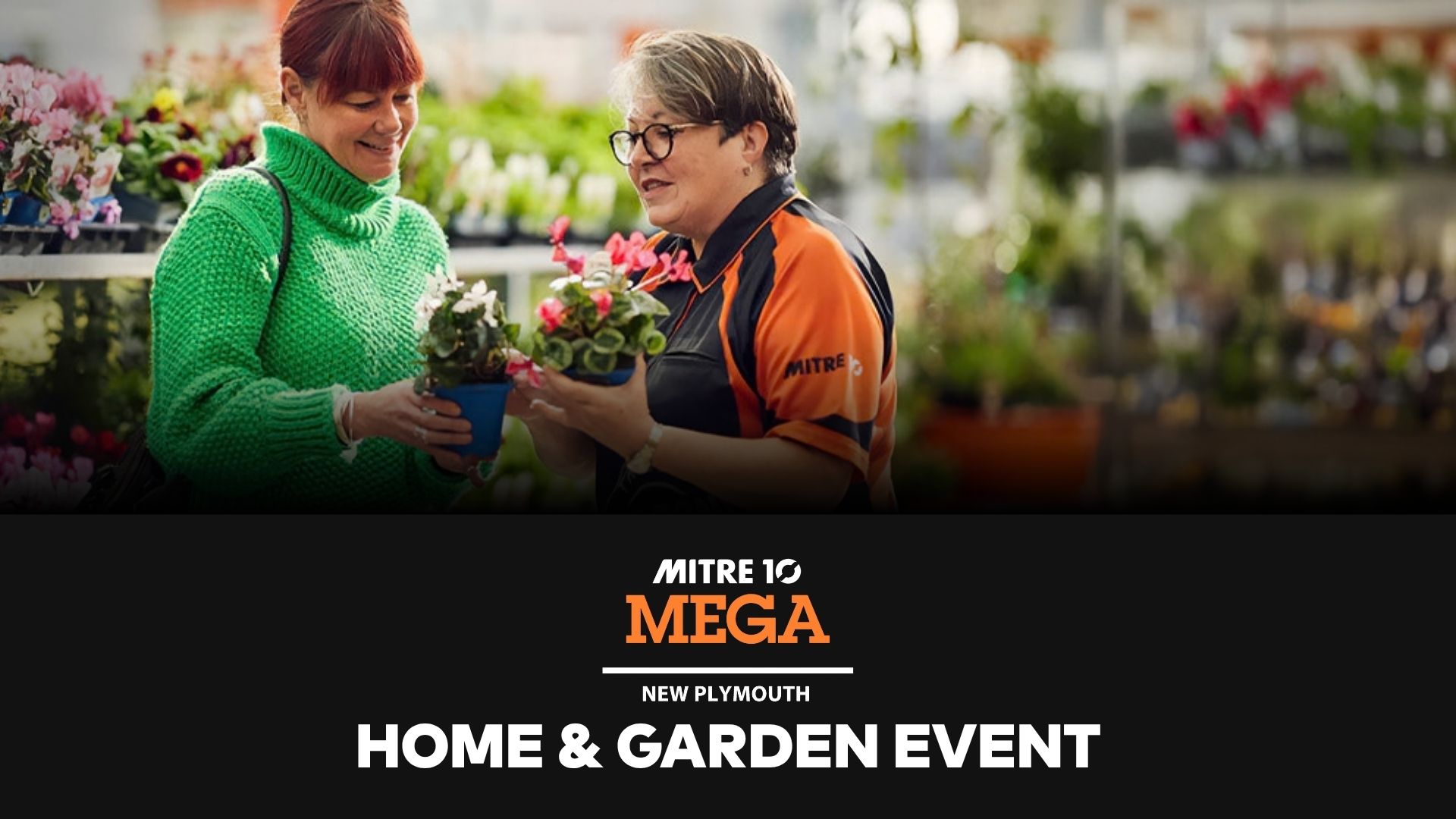 Two smiling women, one in a green sweater and one in a Mitre 10 uniform, are holding potted plants in a garden center.
