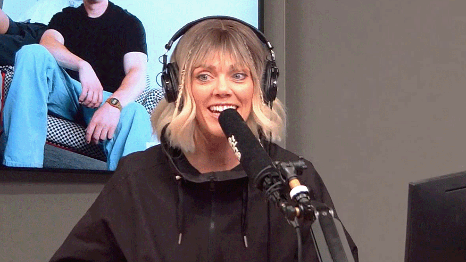 A blonde woman with headphones and a microphone smiles, wearing a black hoodie, with a man in a black t-shirt and blue pants visible on a screen behind her.