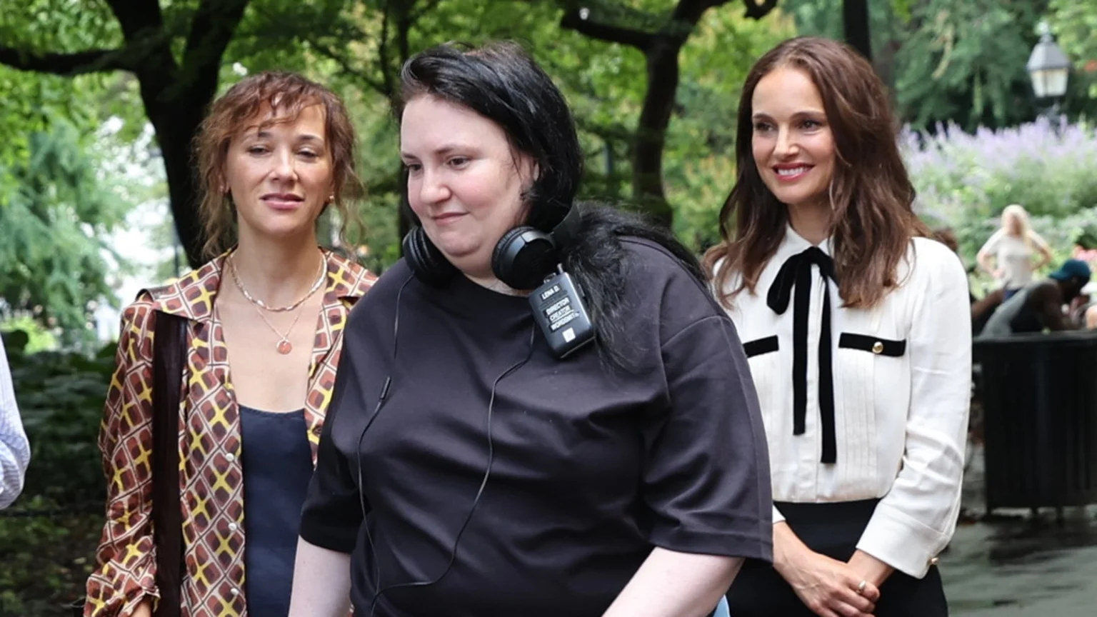 Lena Dunham, wearing headphones and a director badge, stands between a woman in a patterned shirt and Natalie Portman, who smiles in a white blouse with a black tie.