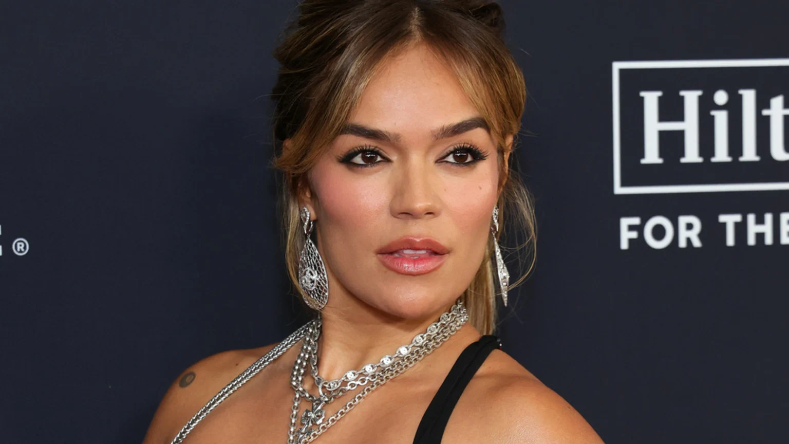 A woman with brown hair, bold makeup, and several silver necklaces, large earrings, and a small tattoo on her shoulder, poses against a dark background with a Hilton logo.