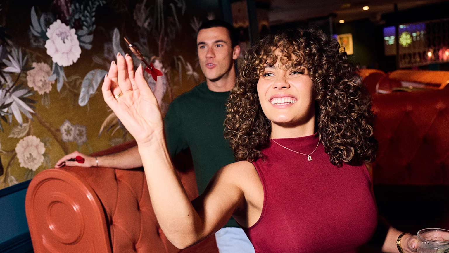 A smiling woman with curly hair appears to throw a dart while a man watches in the background, both in an indoor setting with decorative floral wallpaper and red upholstered seating.