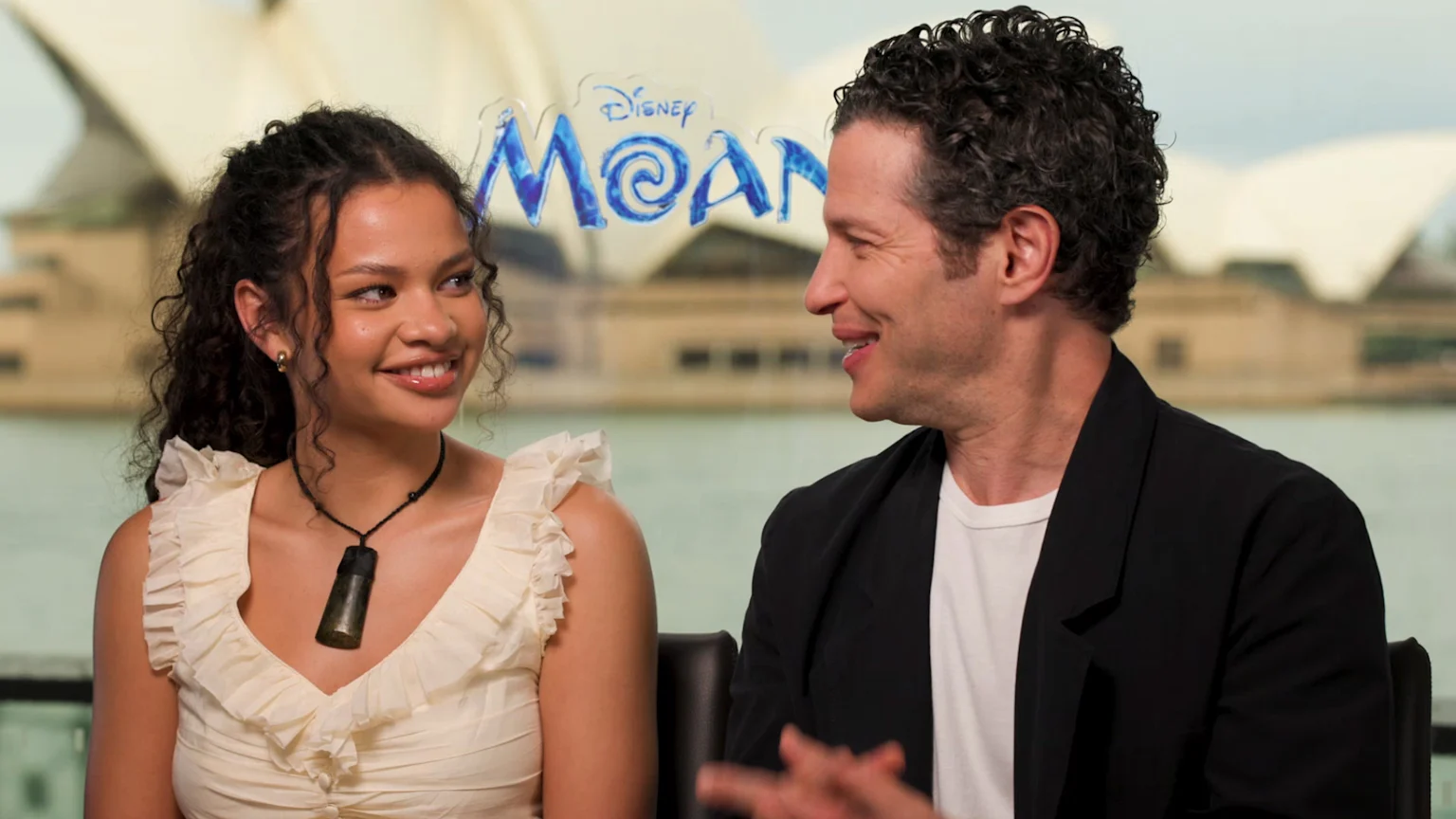 A young woman and a man are smiling and looking at each other, with the Disney Moana logo and the Sydney Opera House visible in the blurred background.