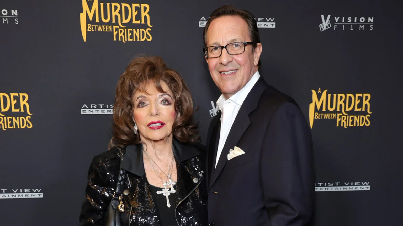 A portrait photograph in an indoor event setting features Joan Collins wearing a black sequined jacket, silver necklaces including a cross pendant, earrings, and carrying a black leather handbag with a gold charm. An unknown man in a dark suit, white shirt, patterned pocket square, and black-framed glasses smiles beside her. They stand before a black fabric promotional backdrop displaying logos for "A Murder Between Friends," Vision Films, and Artist View Entertainment. The lighting is professional and even.