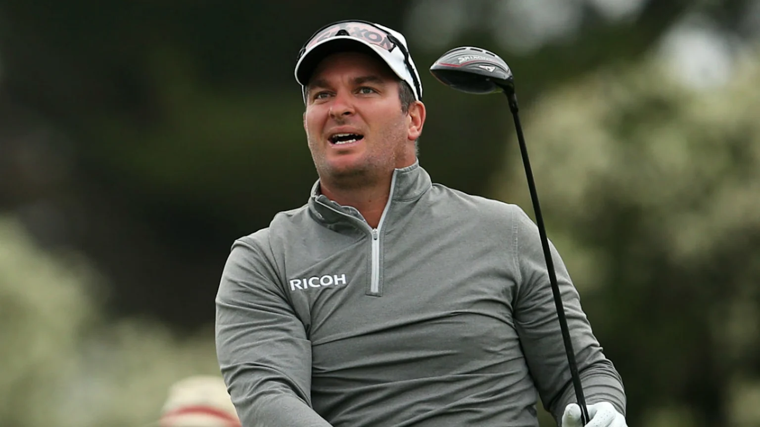 A male golfer in a grey quarter-zip shirt with a RICOH logo, white cap, and sunglasses, holds a golf club and looks with an open mouth against a blurred green background.