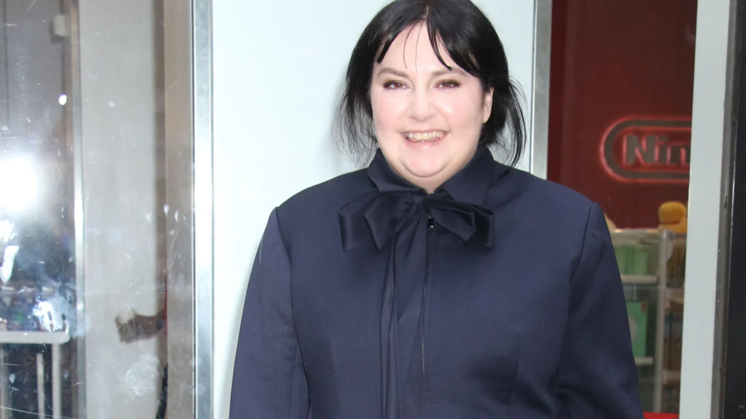 A smiling woman with dark hair and fringe wears a navy top with a bow, standing indoors with a reflection on the left and a blurry red Nintendo logo in the background.