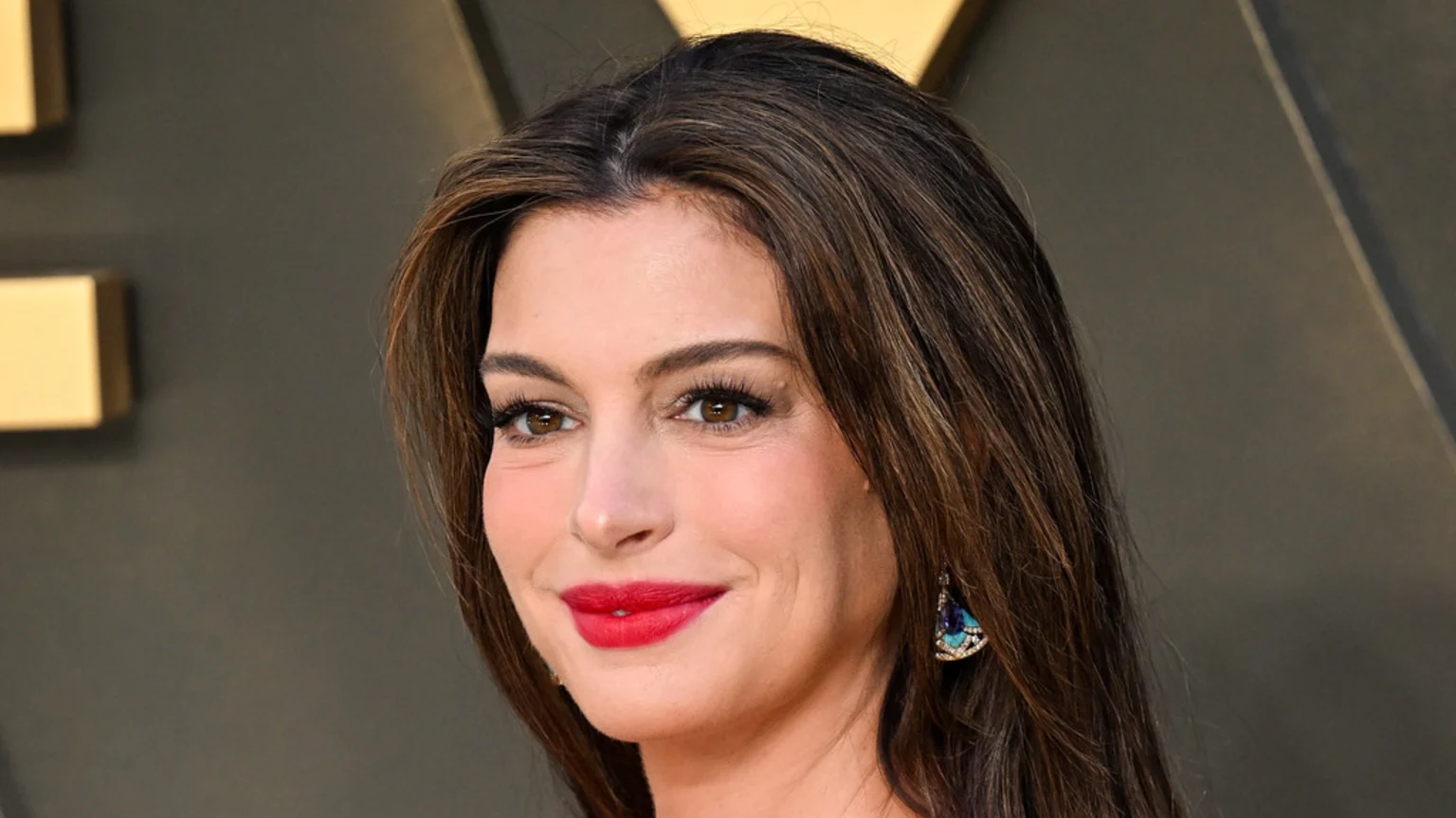 Close-up of Anne Hathaway with long brown hair, red lipstick, a blue and turquoise earring, and a slight smile.