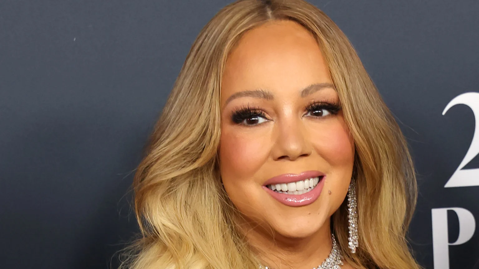 Mariah Carey smiles, head tilted slightly, with long blonde hair, bold eye makeup, glossy pink lips, and shimmering diamond earrings and necklace.