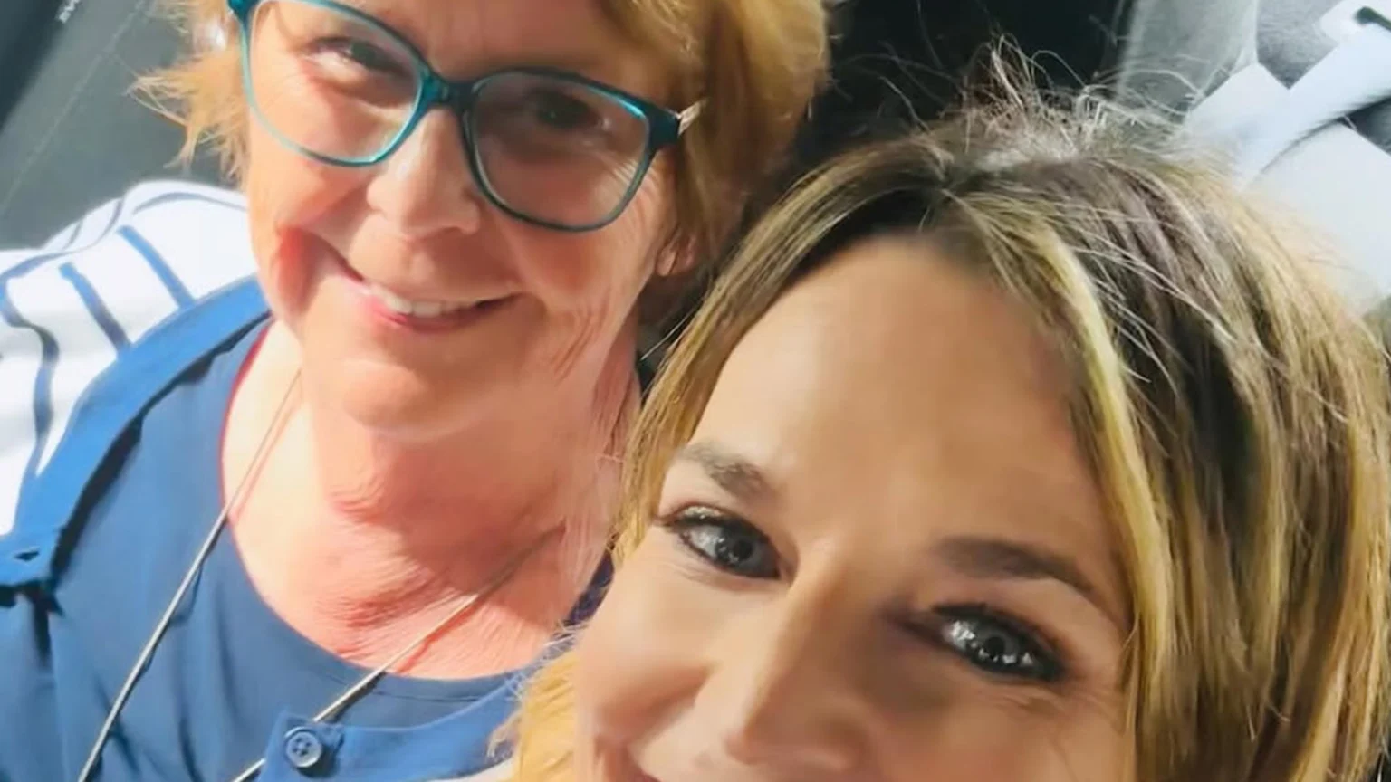 A bright indoor selfie showing two women: an older woman with teal glasses, blue v-neck top, and striped cardigan smiling broadly, and a younger blonde woman with makeup in the foreground also smiling.