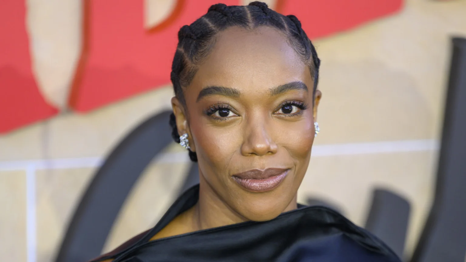 A close-up headshot of actress Naomi Ackie smiling at the camera, featuring her braided hair, sparkling earrings, and a dark, silky outfit against a blurred red and beige background.