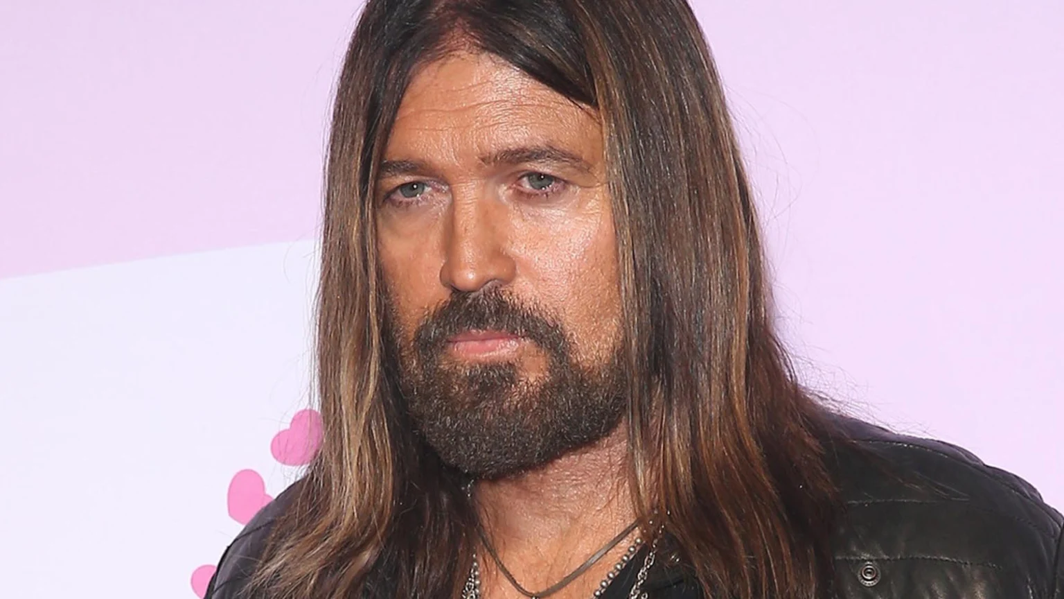 Close-up of Billy Ray Cyrus, a man with long brown hair and a beard, looking forward, wearing a black leather jacket and necklaces against a pink and white background.