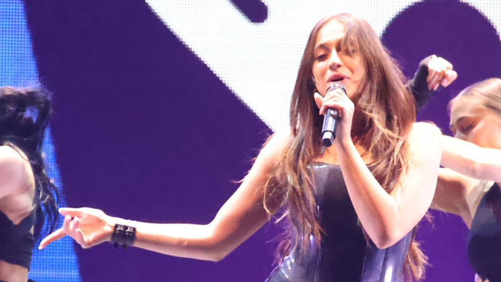 A female singer with long brown hair performs on stage, holding a microphone to her mouth, wearing a dark top, with her left arm extended and other performers visible in the background.