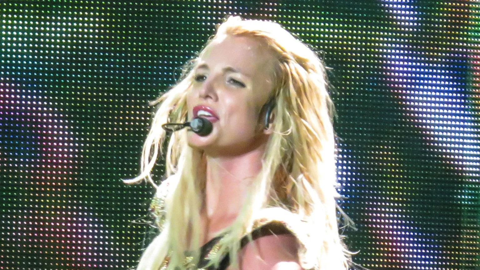 Britney Spears performs on stage, wearing a headset microphone, with her blonde hair backlit by a vibrant, colorful LED screen.
