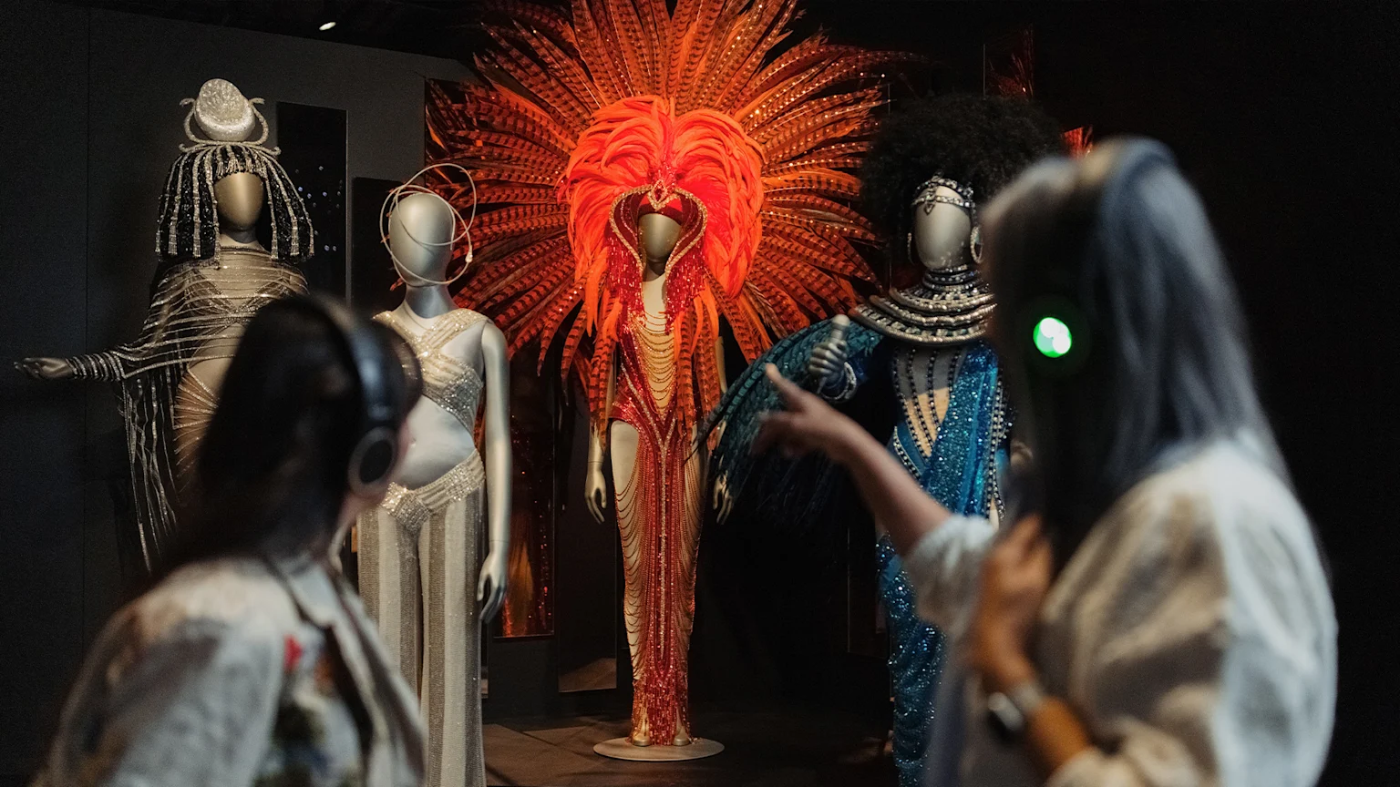 Two women wearing headphones, back facing the camera, turned and facing an extravagant orange outfit with a feather headpiece.