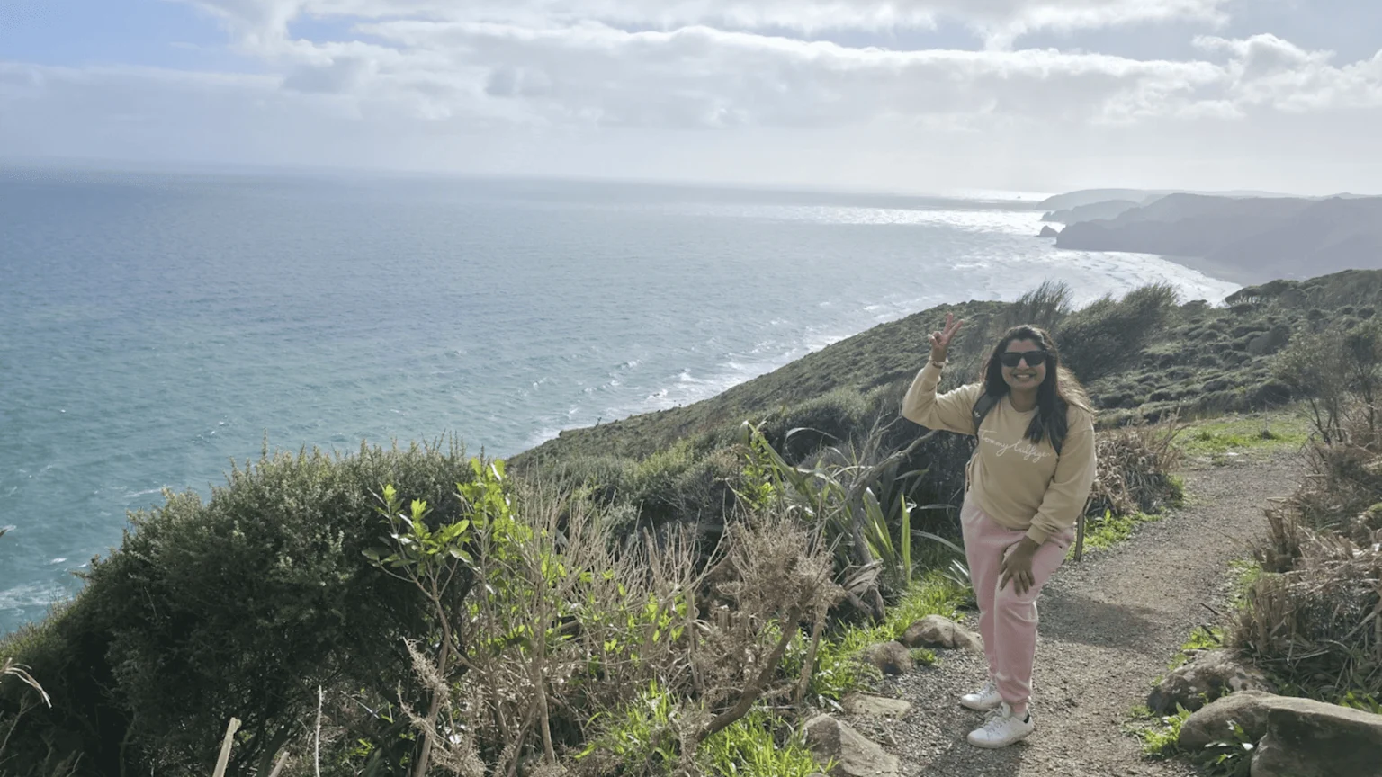 A woman in sunglasses and casual clothes smiles and makes a peace sign on a coastal hiking path, overlooking a vast ocean and distant cliffs.