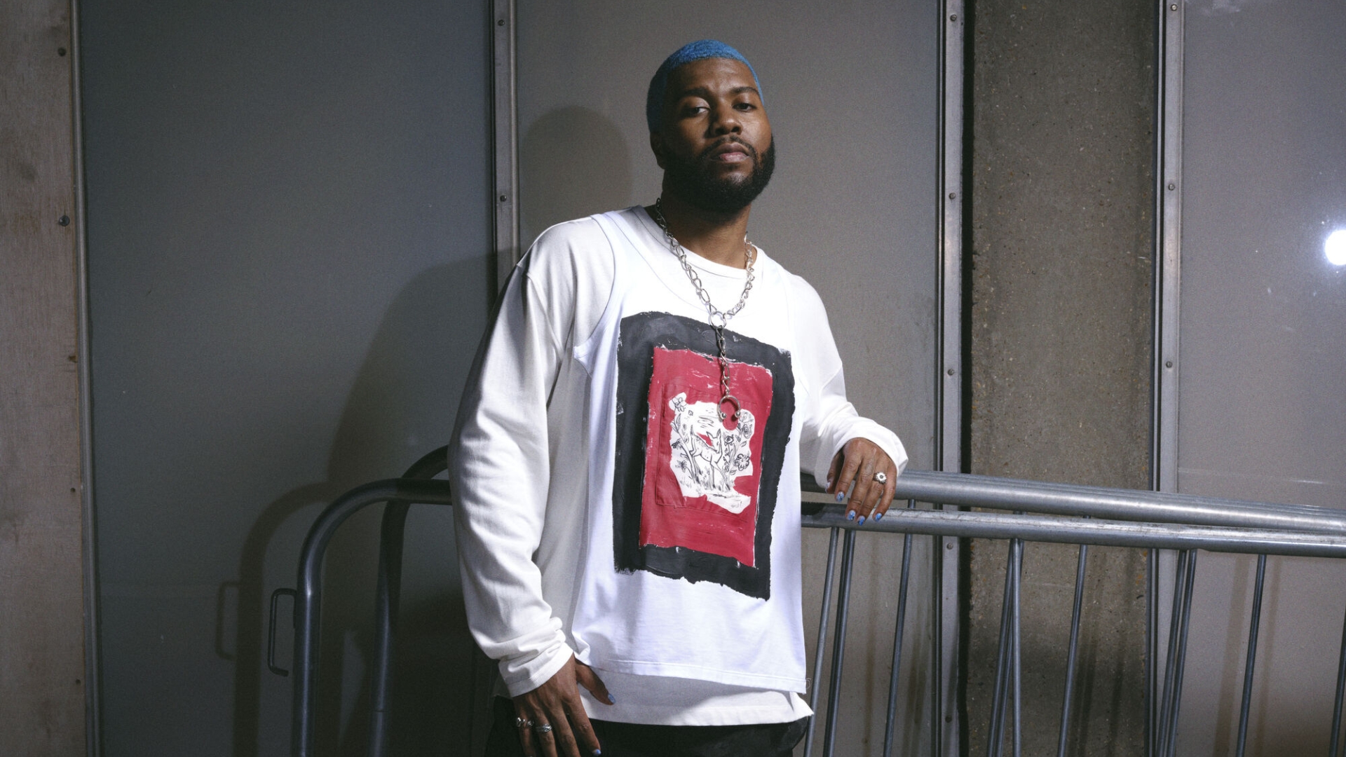 A man with blue hair and a beard, wearing a white long-sleeved shirt with a black and red graphic print, a silver chain, rings, and blue nail polish, leans on a metal barrier.