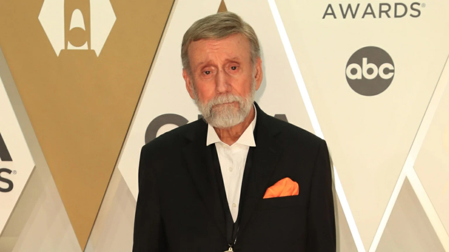 Country singer Ray Stevens, an elderly man with a grey beard and hair, wears a black suit and an orange pocket square at an event.