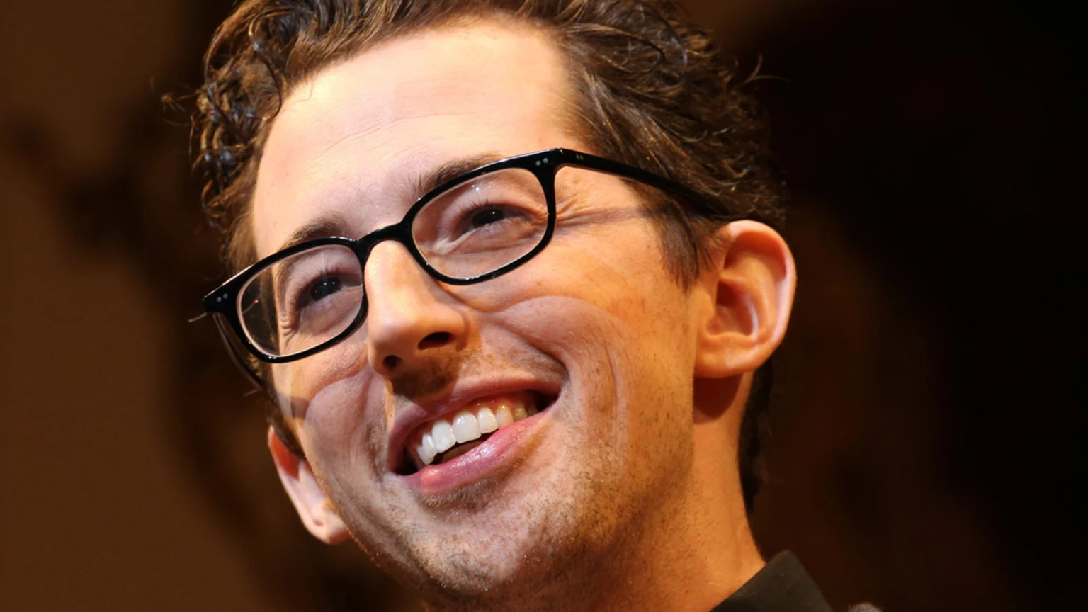 A close-up portrait of a smiling man with dark, wavy hair and black-framed glasses, looking upwards with a broad grin showing his white teeth.