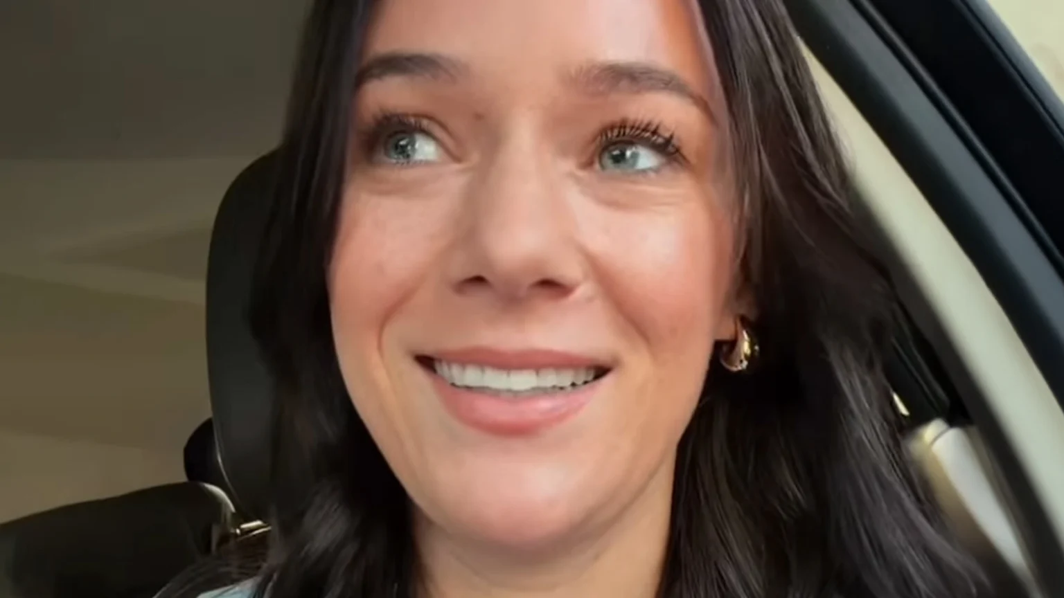 A smiling woman with dark hair and bright blue eyes looks up and to her left while sitting in a car, wearing a small gold hoop earring.
