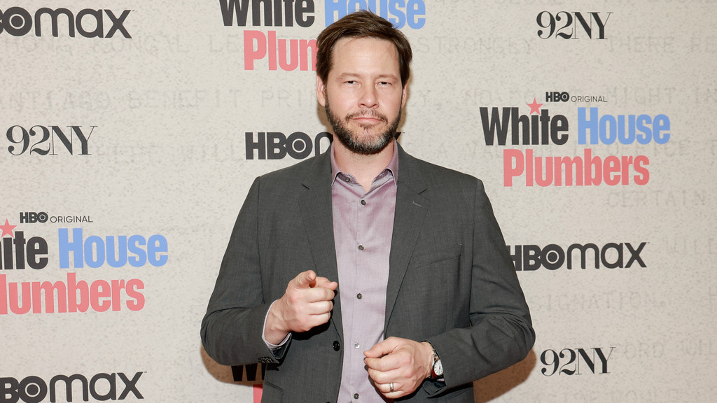 A bearded man in a grey suit jacket and purple collared shirt stands against a promotional backdrop for 'White House Plumbers', pointing with his right hand.