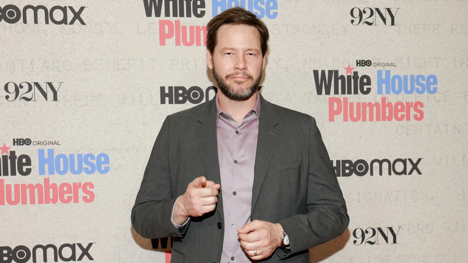 A bearded man in a grey suit jacket and purple collared shirt stands against a promotional backdrop for 'White House Plumbers', pointing with his right hand.