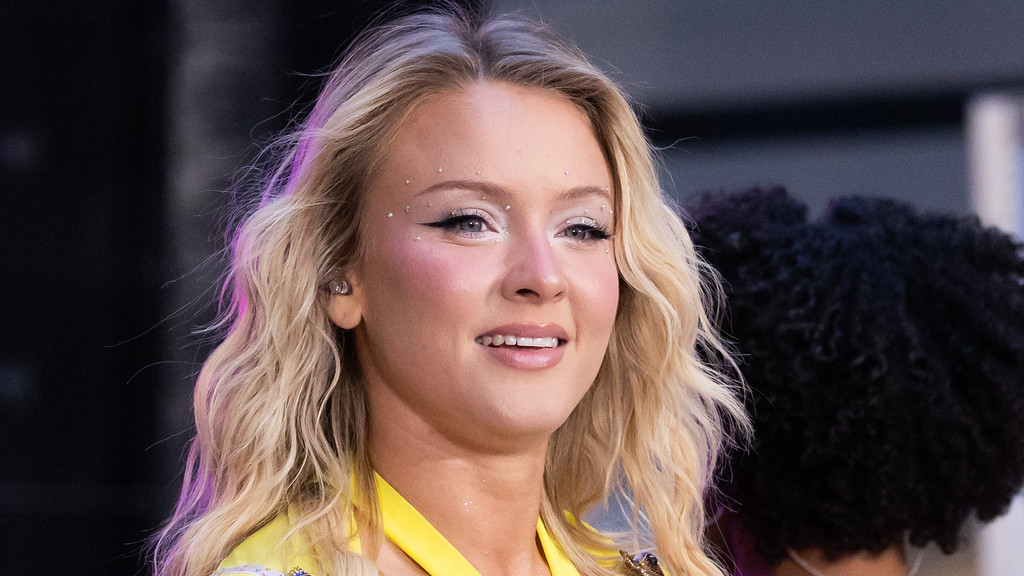 A young woman with wavy blonde hair and a subtle smile wears a yellow outfit, featuring elaborate eye makeup with winged eyeliner, glitter, and rhinestones.