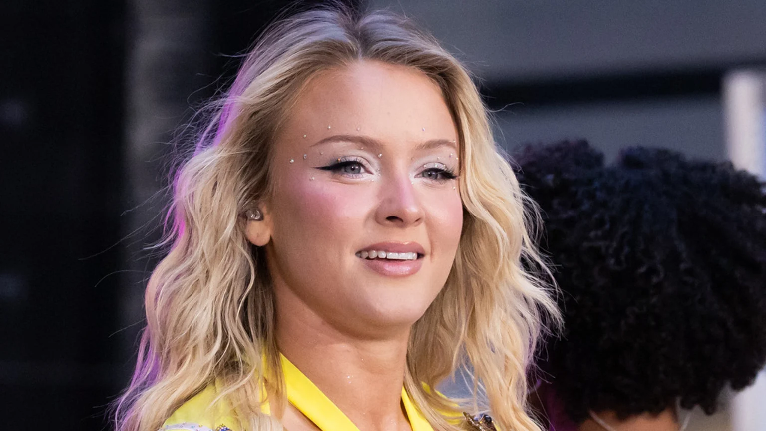A young woman with wavy blonde hair and a subtle smile wears a yellow outfit, featuring elaborate eye makeup with winged eyeliner, glitter, and rhinestones.