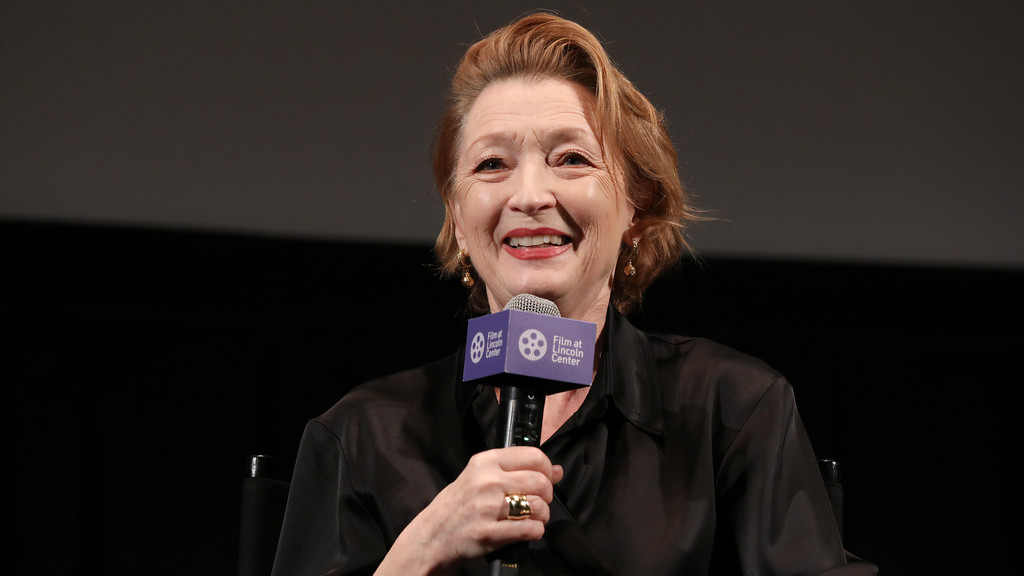 A smiling woman with auburn hair holds a microphone displaying the 'Film at Lincoln Center' logo, against a dark background.