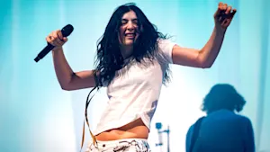 A mid-shot captures Lorde singing passionately into a microphone on stage, wearing a white t-shirt and metallic-accented pants. Bright blue and white stage lights illuminate the scene with another blurred figure in the background.