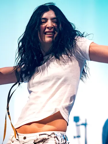 A mid-shot captures Lorde singing passionately into a microphone on stage, wearing a white t-shirt and metallic-accented pants. Bright blue and white stage lights illuminate the scene with another blurred figure in the background.