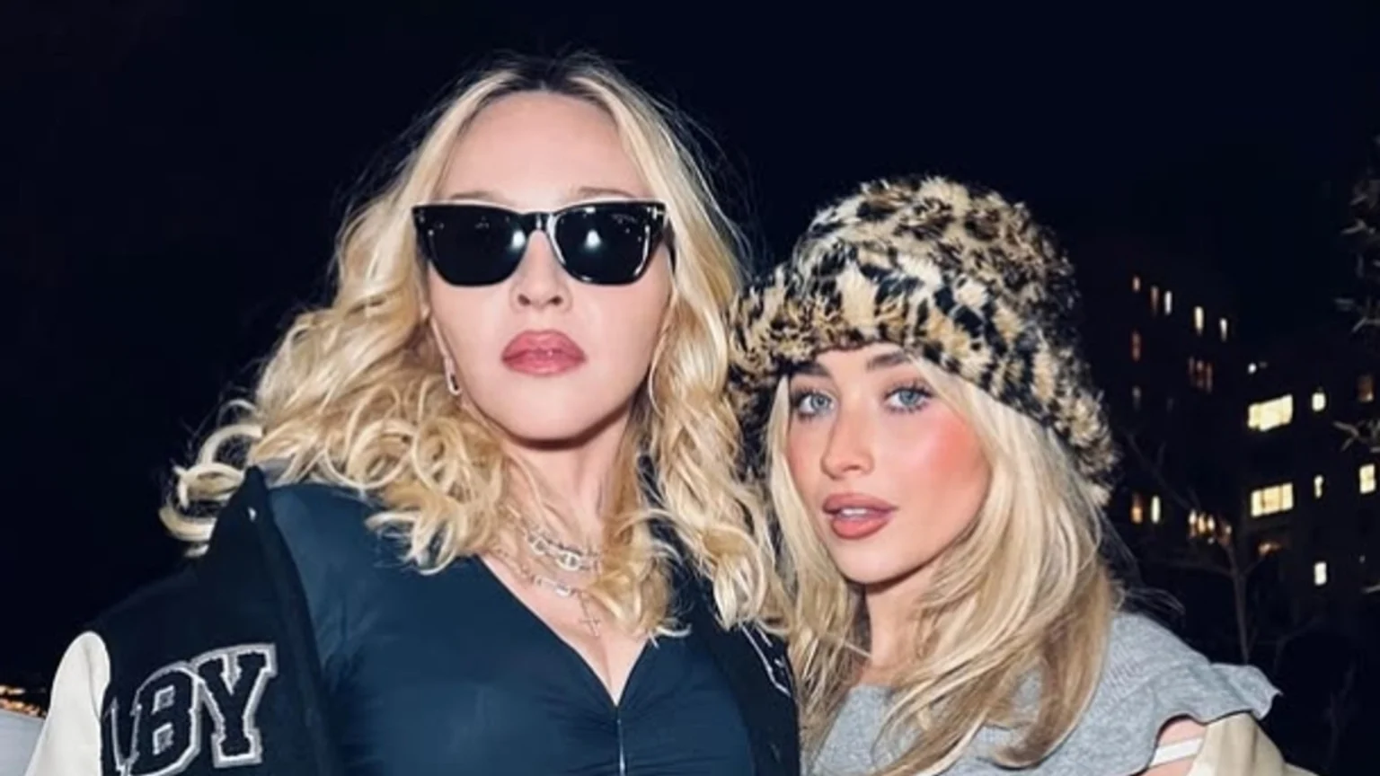 Madonna, wearing sunglasses, a black top, and a varsity jacket, poses next to Sabrina Carpenter, who wears a leopard print hat and a grey top, both looking towards the camera at night.