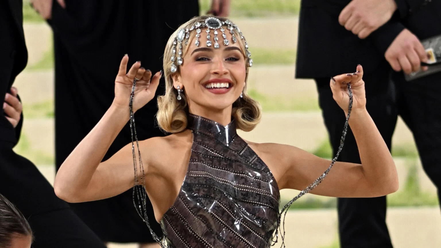 A smiling woman with blonde hair in retro curls and a jeweled headpiece wears a unique halter-neck dress resembling film strips, holding dark beaded strands in her raised hands.