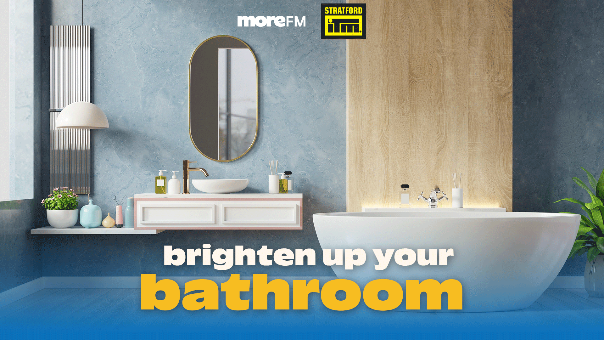 A modern bathroom with a white freestanding tub, floating vanity, gold faucet, oval mirror, light blue walls, and wood paneling, encouraging to 'brighten up your bathroom' with MoreFM and Stratford IM logos.