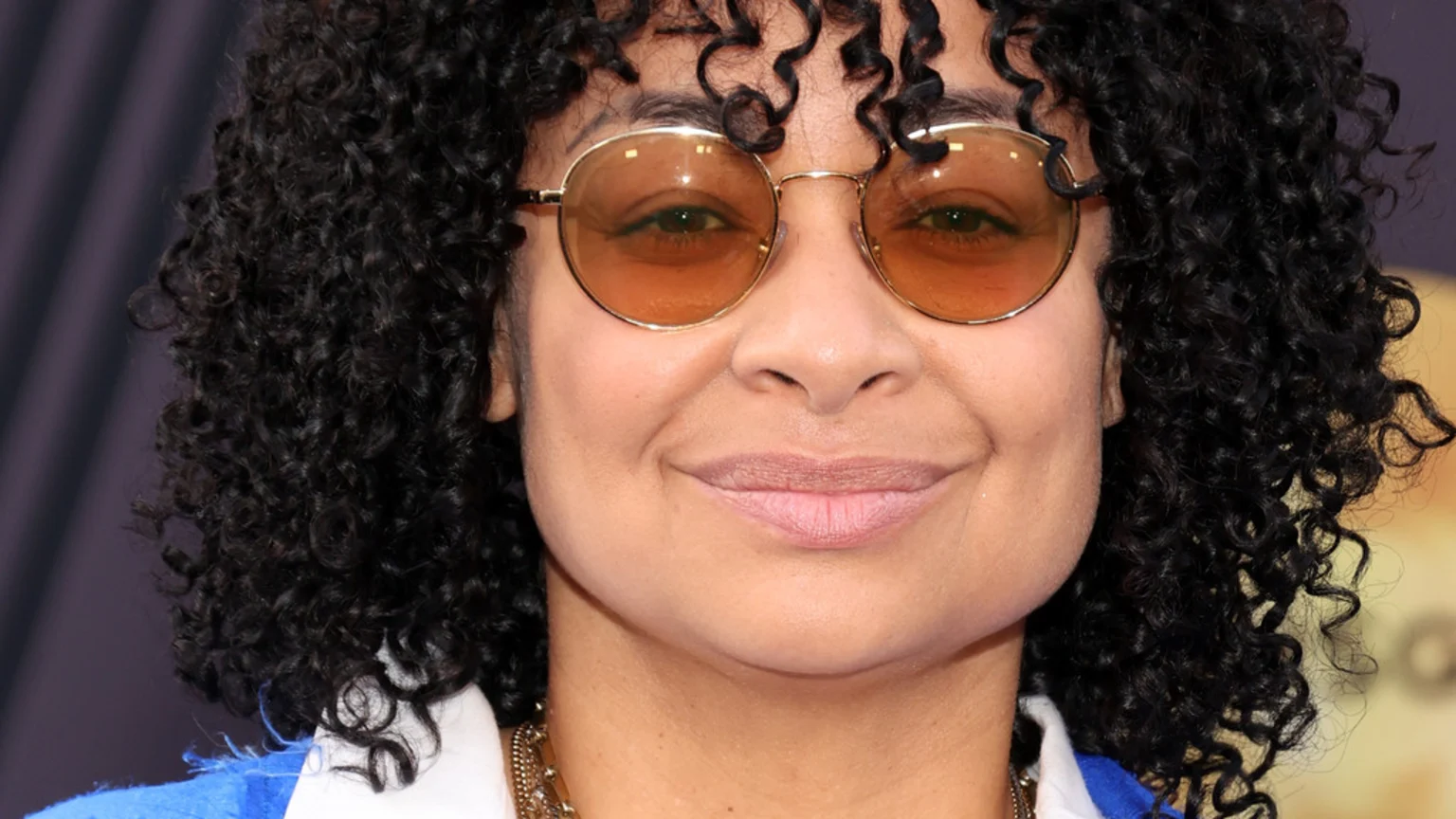 A close-up of a smiling person with dark curly hair, wearing round, amber-tinted sunglasses, a white collar, and a gold chain necklace.