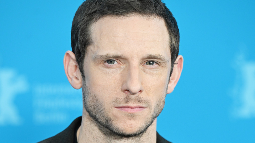 Close-up portrait of a man with short dark hair, green eyes, light stubble, and a neutral expression, against a blue background.
