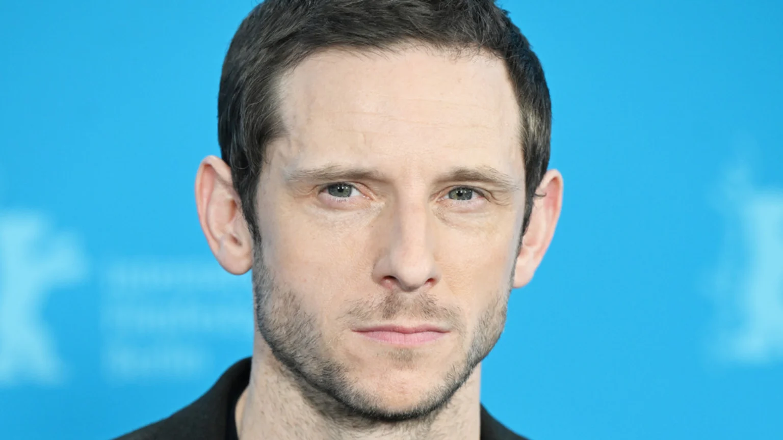 Close-up portrait of a man with short dark hair, green eyes, light stubble, and a neutral expression, against a blue background.