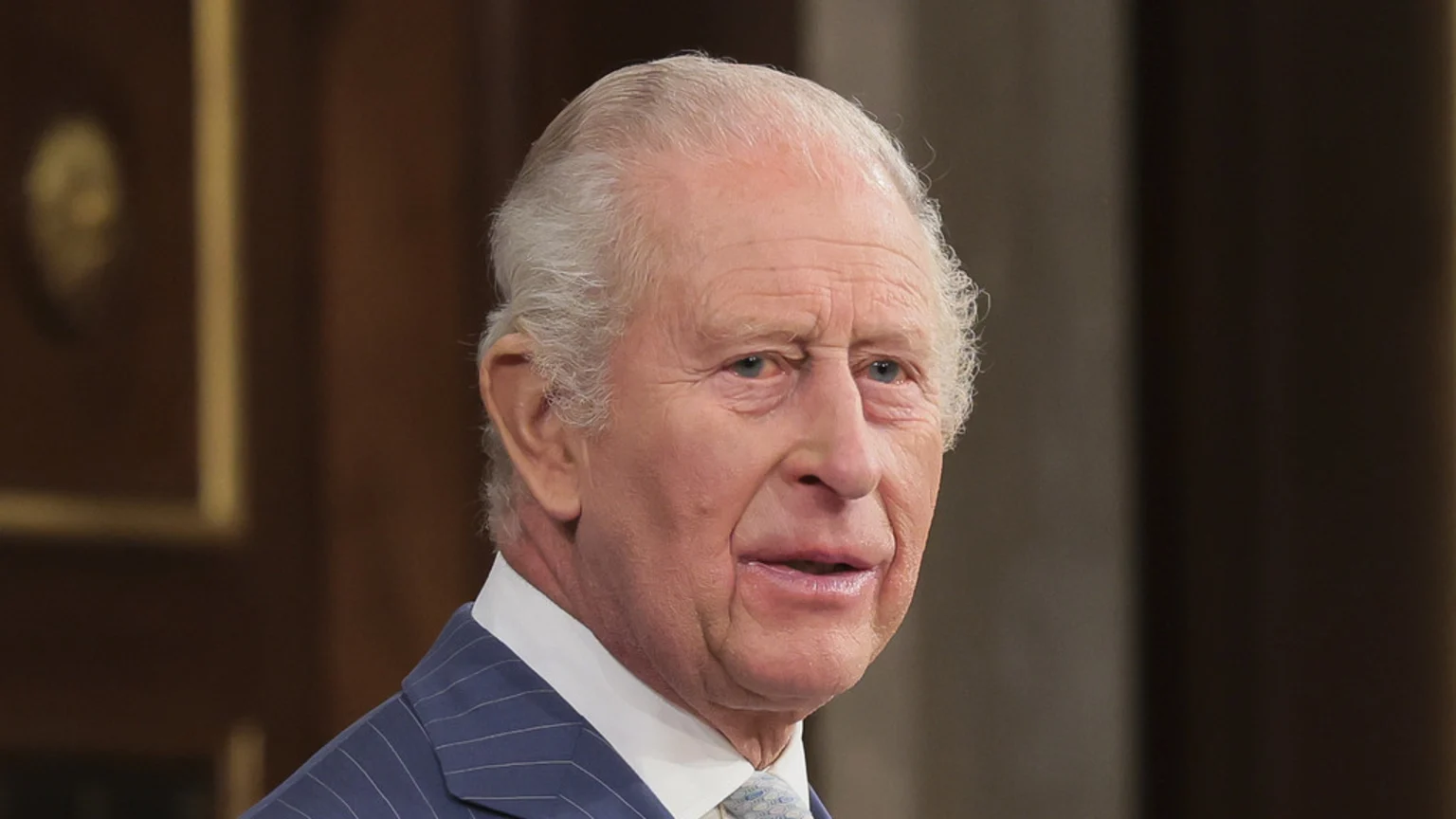 A close-up shot of King Charles III, an older man with white hair, wearing a pinstriped suit, white shirt, and light tie, looking towards the right with a neutral expression.
