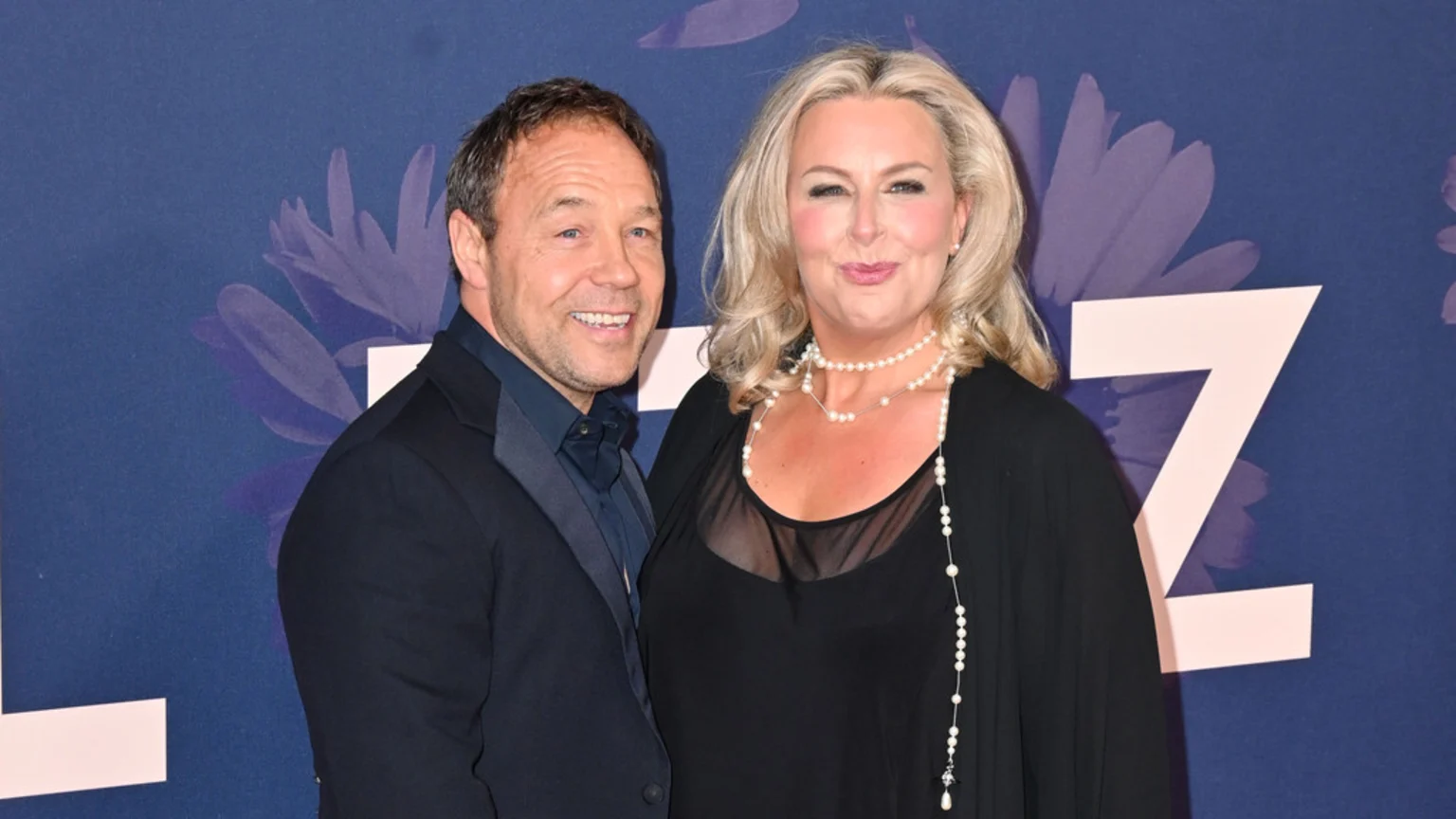 A man with short brown hair in a dark suit smiles next to a woman with blonde hair in a black top and pearl necklaces, both standing against a blue floral background.