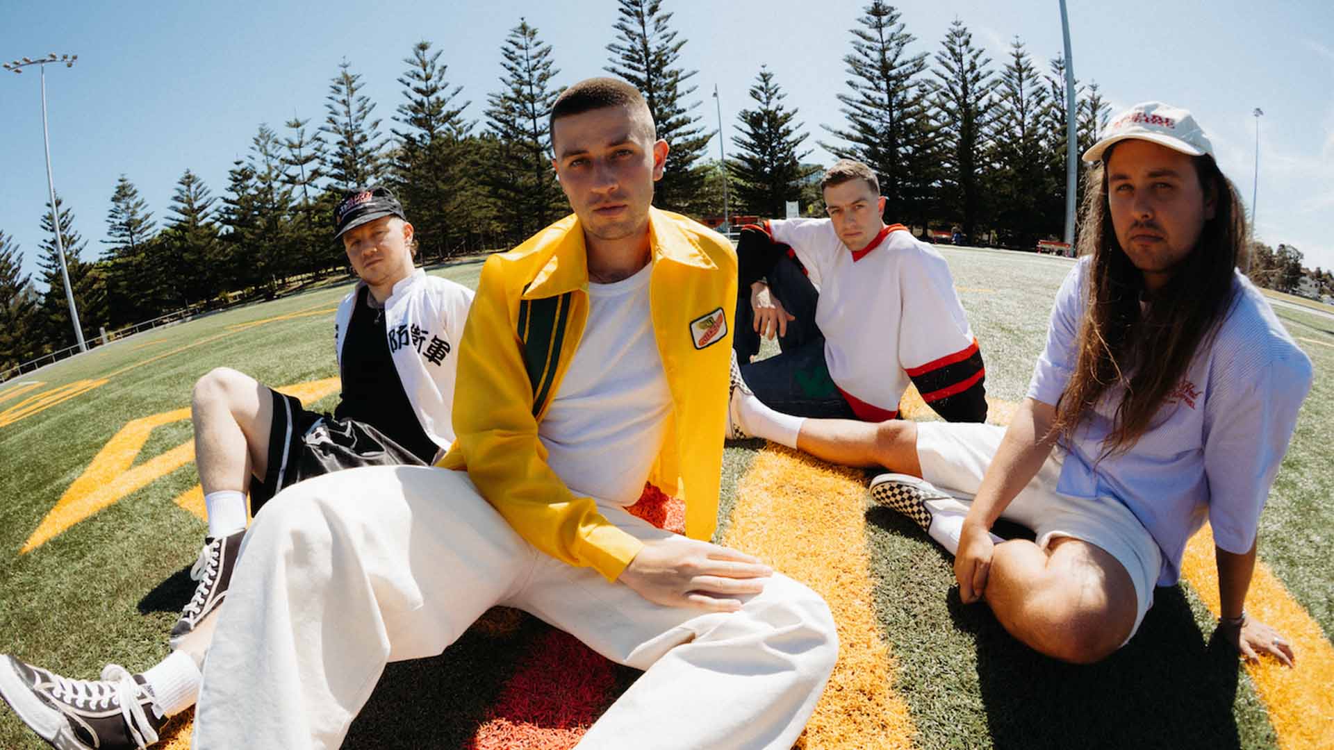 Four men sit on a green sports field with yellow and red markings under a clear sky, wearing casual outfits, with pine trees in the background.