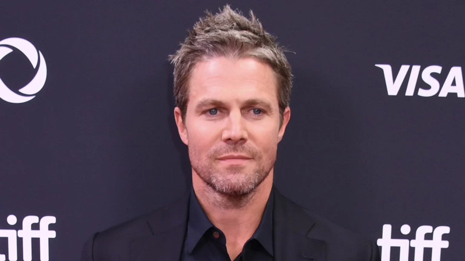 Stephen Amell, a man with graying hair and a beard, looks forward while wearing a dark suit against a dark background with sponsor logos.