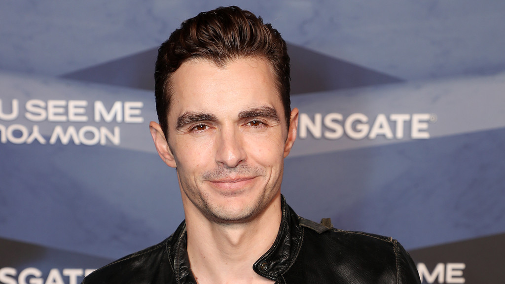 Close-up of Dave Franco, smiling subtly, with dark hair and a black leather jacket, against a blurry background with "YOU SEE ME" and "LIONSGATE" text.