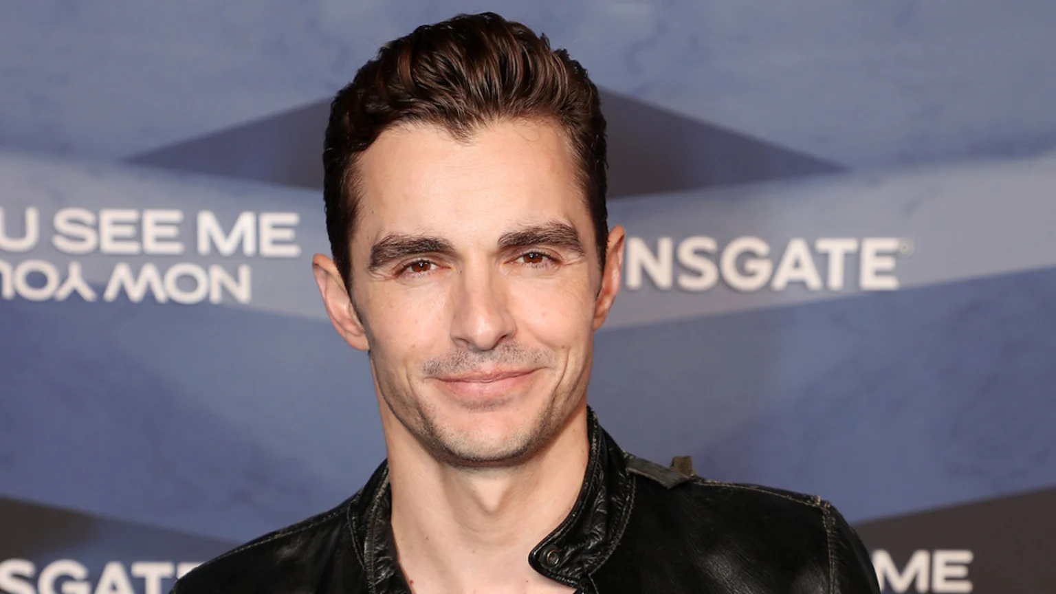 Close-up of Dave Franco, smiling subtly, with dark hair and a black leather jacket, against a blurry background with "YOU SEE ME" and "LIONSGATE" text.