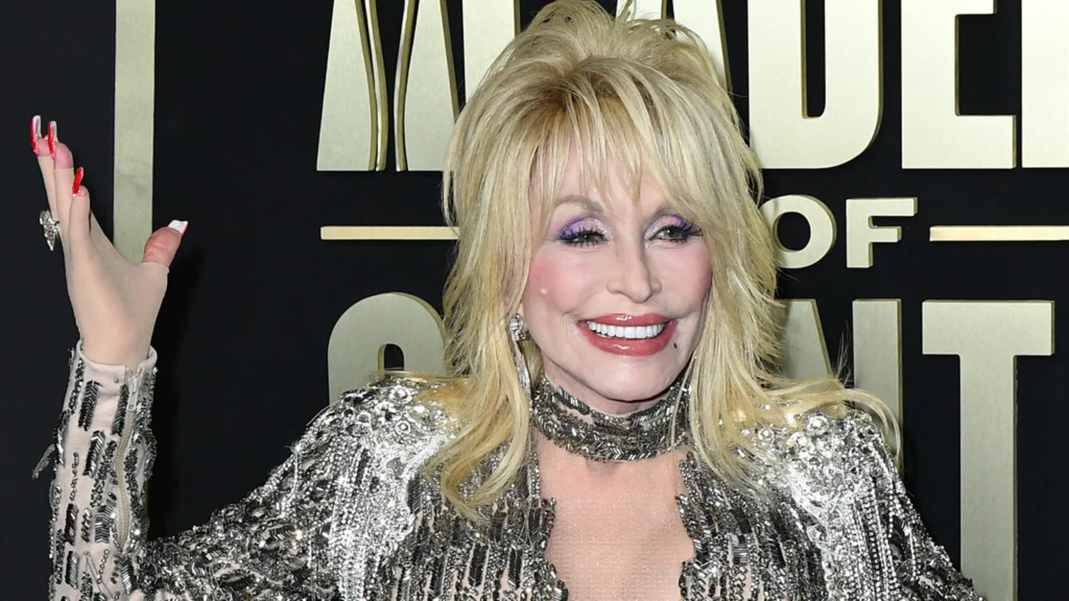 Dolly Parton smiles broadly, wearing a sparkly silver outfit, a metallic choker, purple eyeshadow, and long red and white nails on her raised hand, against a black and gold background.