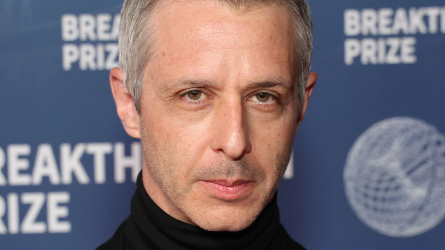 A man with short grey hair, light eyes, and light stubble, wearing a black turtleneck, looks directly at the camera against a blurry blue "BREAKTHROUGH PRIZE" background.