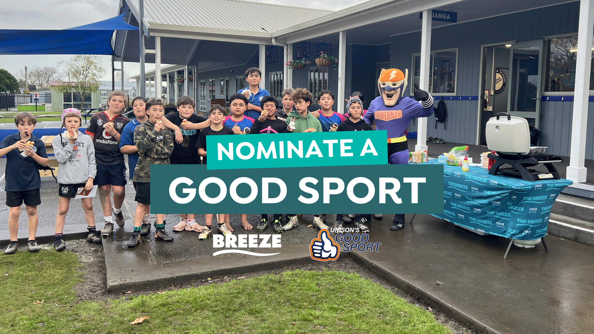 A superhero mascot stands with a group of boys eating hot dogs next to a branded table with a grill, all beneath an overlay saying "NOMINATE A GOOD SPORT".