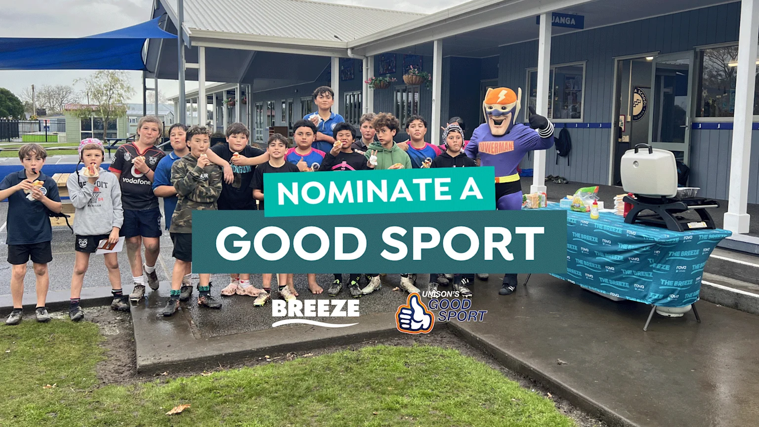 A superhero mascot stands with a group of boys eating hot dogs next to a branded table with a grill, all beneath an overlay saying "NOMINATE A GOOD SPORT".